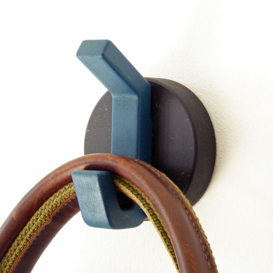 A contemporary wooden robe hook with a blue hook on a black round backplate, mounted on a wall holding a tan leather bag.