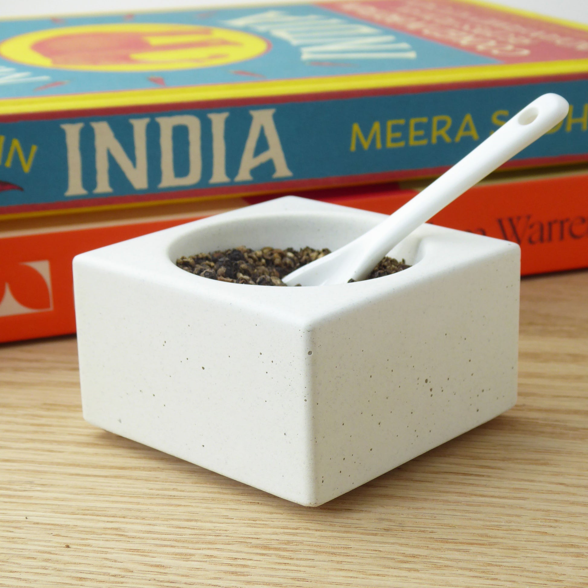 White concrete bowl holding back peppercorns with a white ceramic spoon on a wooden table with colourful cookbooks in the background