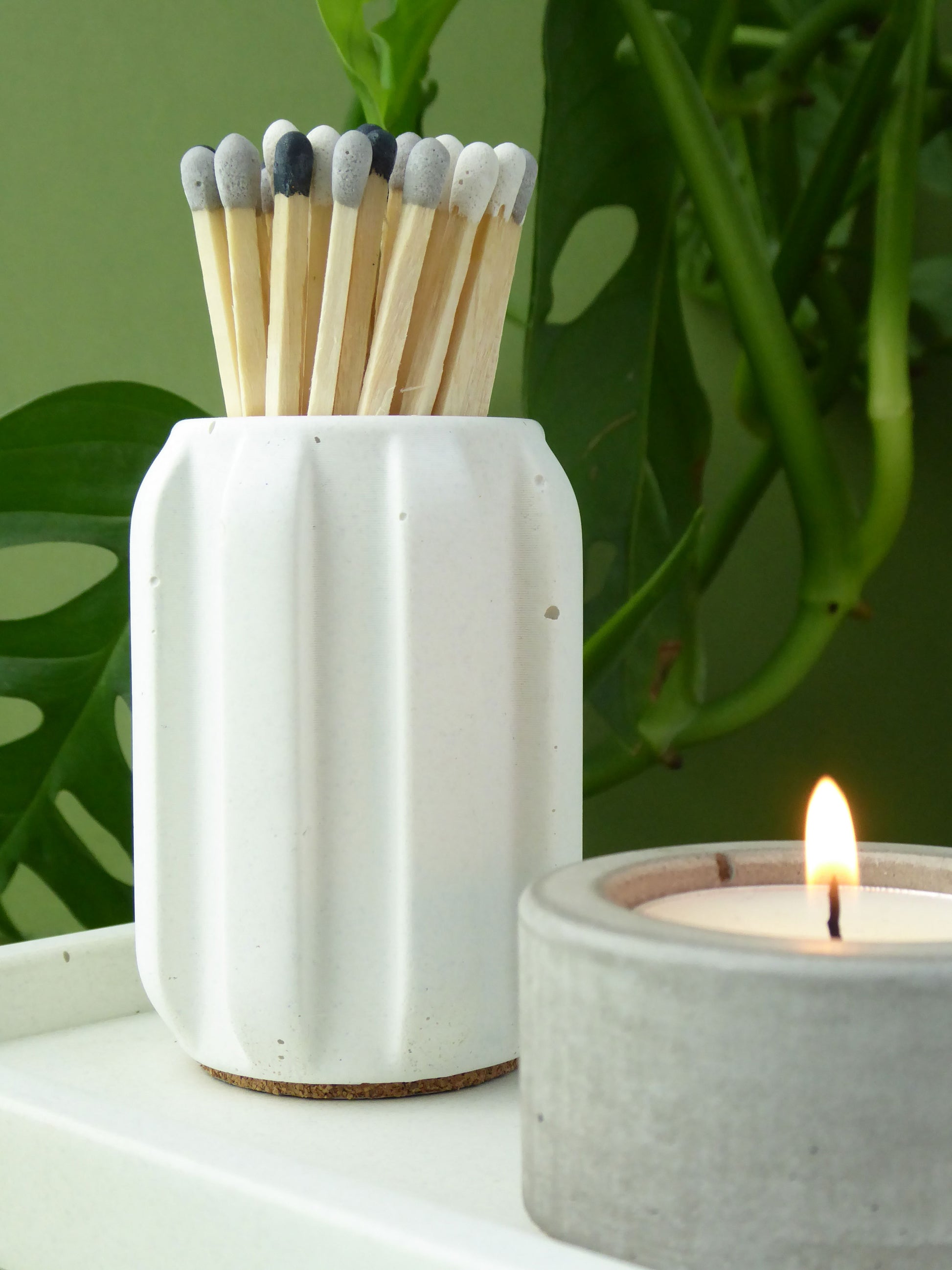 Modern white concrete match holder with long coloured matches next to a lit candle on a green leafy background