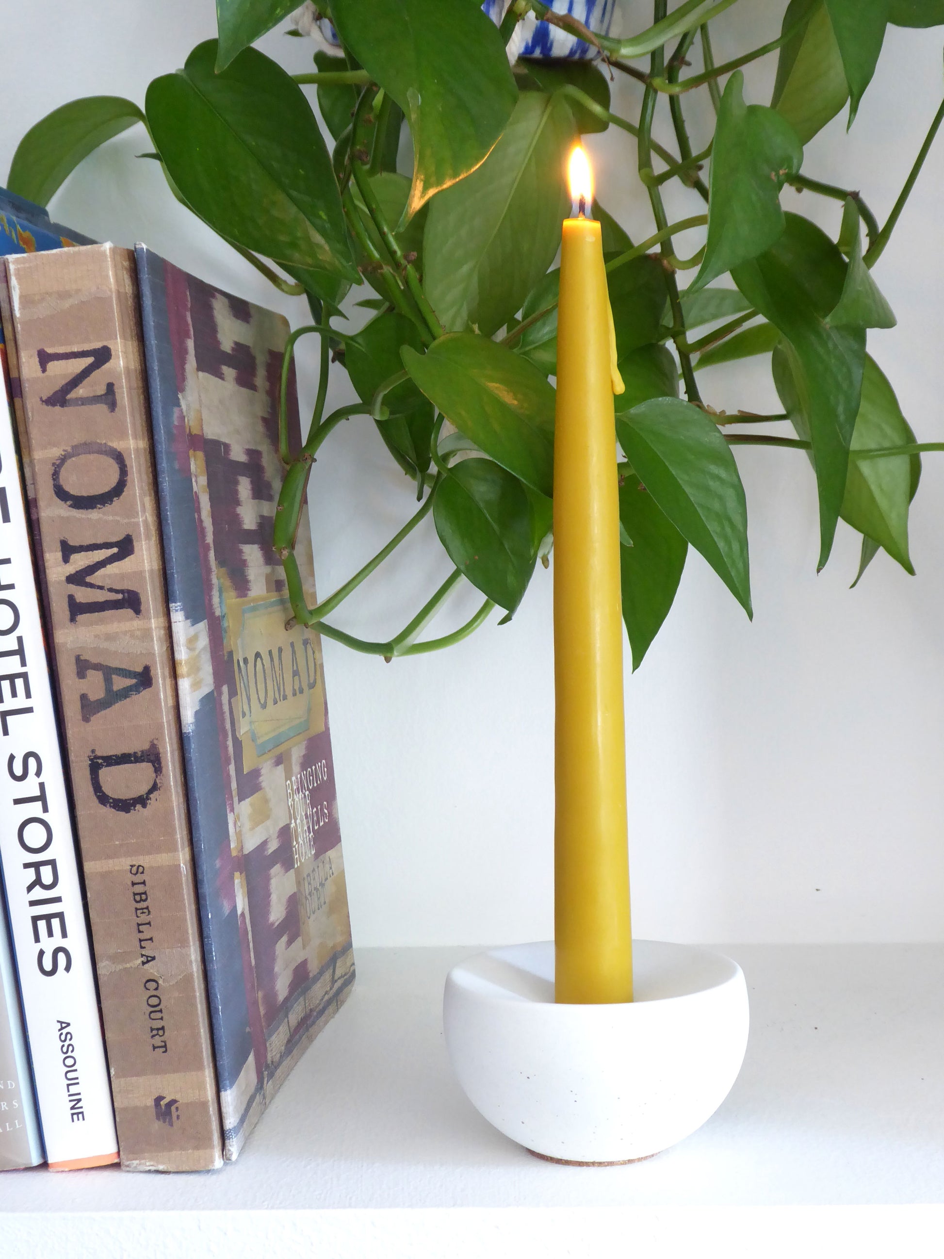 Yellow candle in a white concrete candle holder on a shelf with books and a plant in the background
