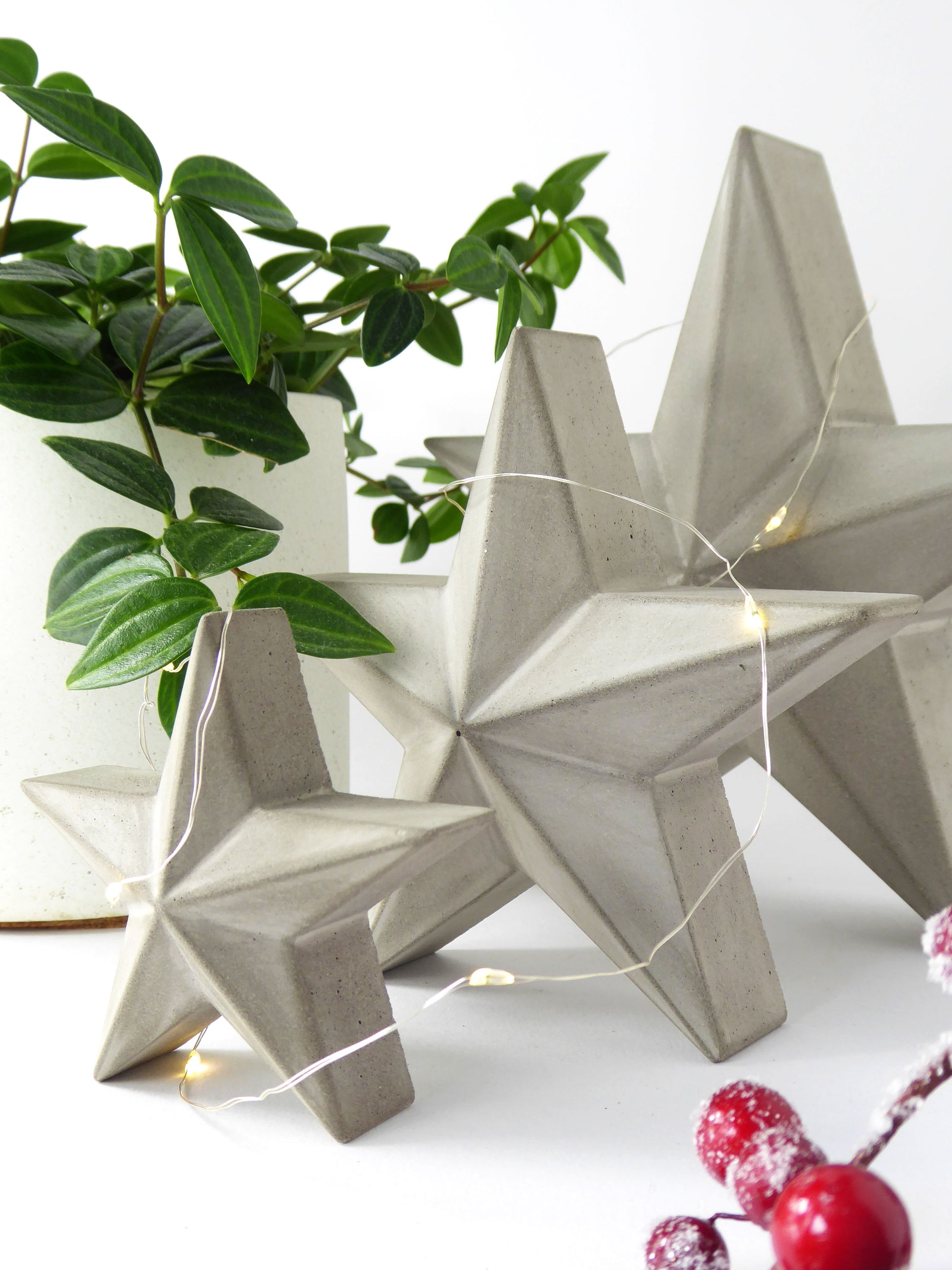 Set of 3 geometric concrete star-shaped freestanding decorations with festive string lights on a white surface with greenery.