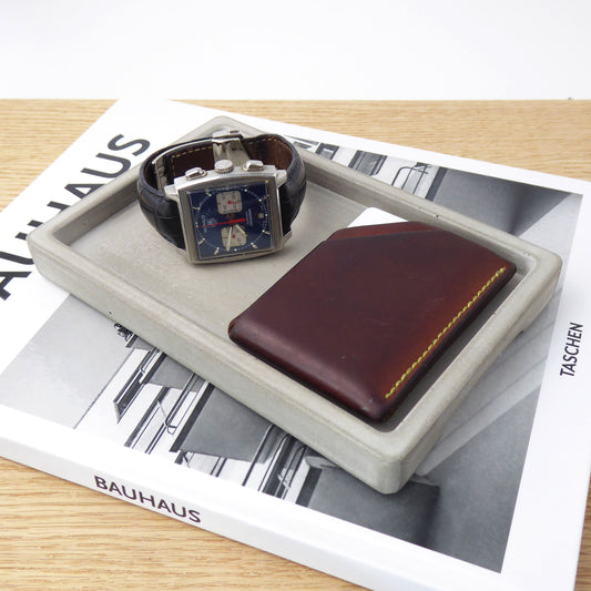 Grey concrete rectangular tray which holds a mens wristwatch and leather wallet on a bedside table