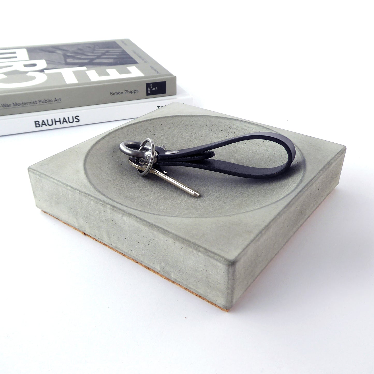 A square-shaped, chunky concrete tray in a natural grey finish holding keys with books in the background