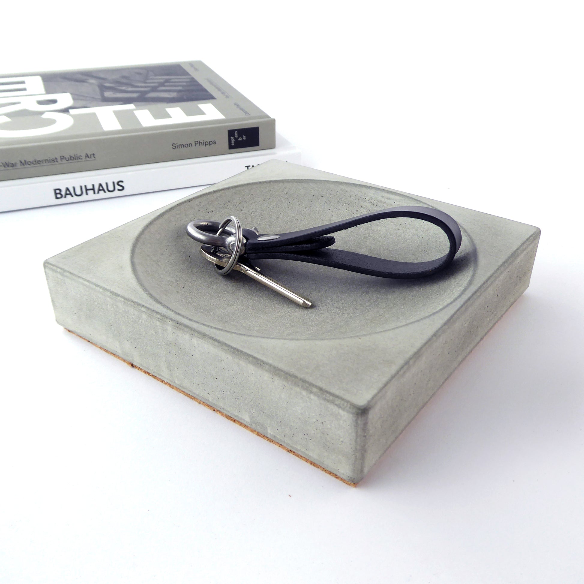 A square-shaped, chunky concrete tray in a natural grey finish holding keys with books in the background