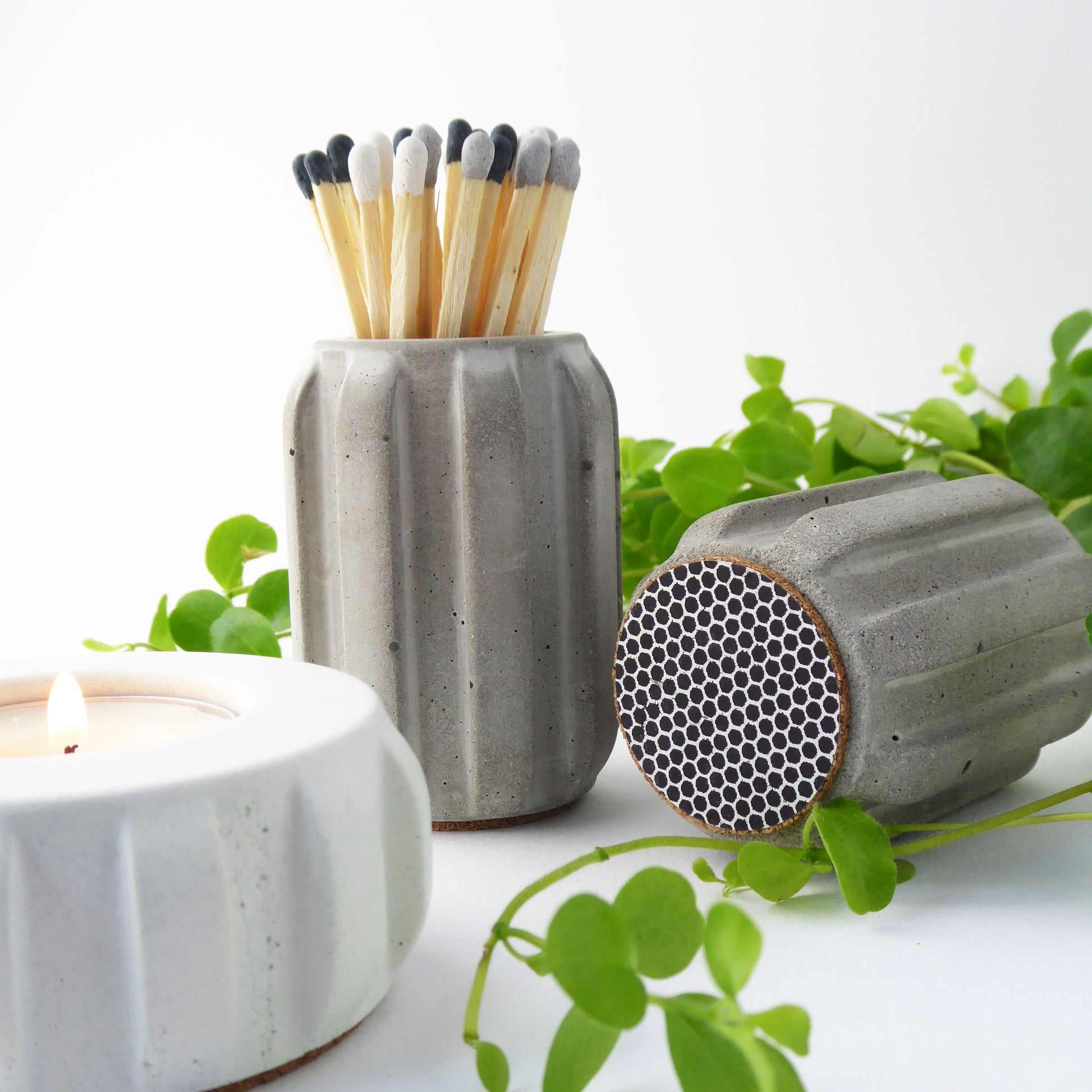 Grey concrete match pot with a ribbed design holding long coloured matches, together with a white concrete candle, and another match pot showing the match striker on the base of the pot.