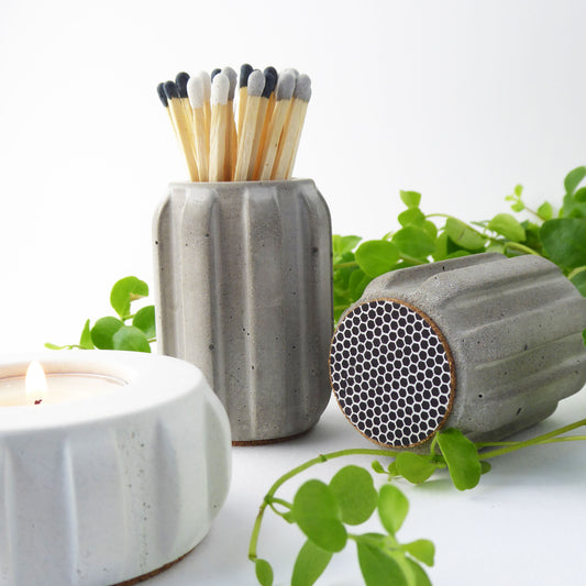 Grey concrete match pot with a ribbed design holding long coloured matches, together with a white concrete candle, and another match pot showing the match striker on the base of the pot.