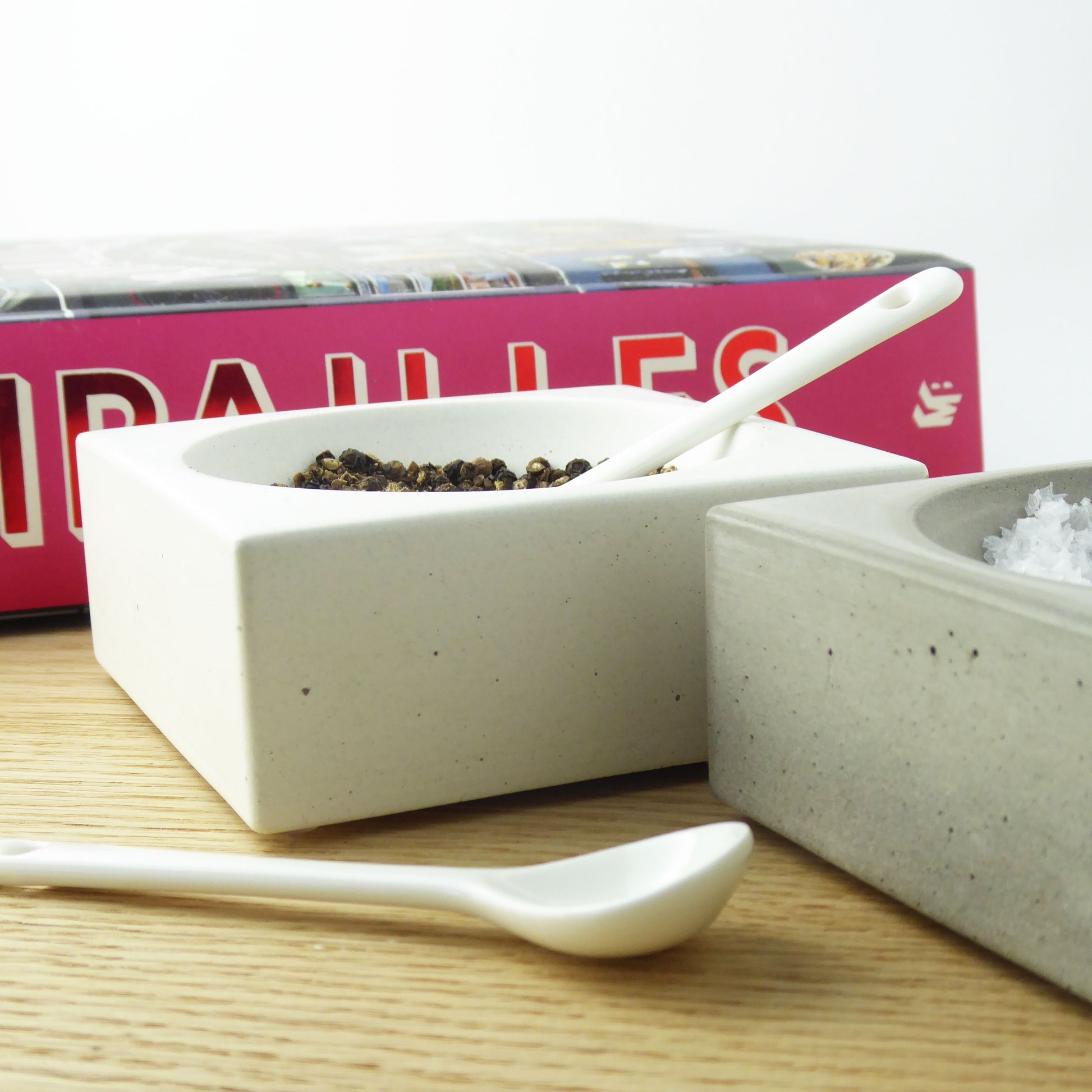Two concrete bowls containing seasalt and black peppercorns with white spoons on a wooden dining table  with a pink cookbook in the background.