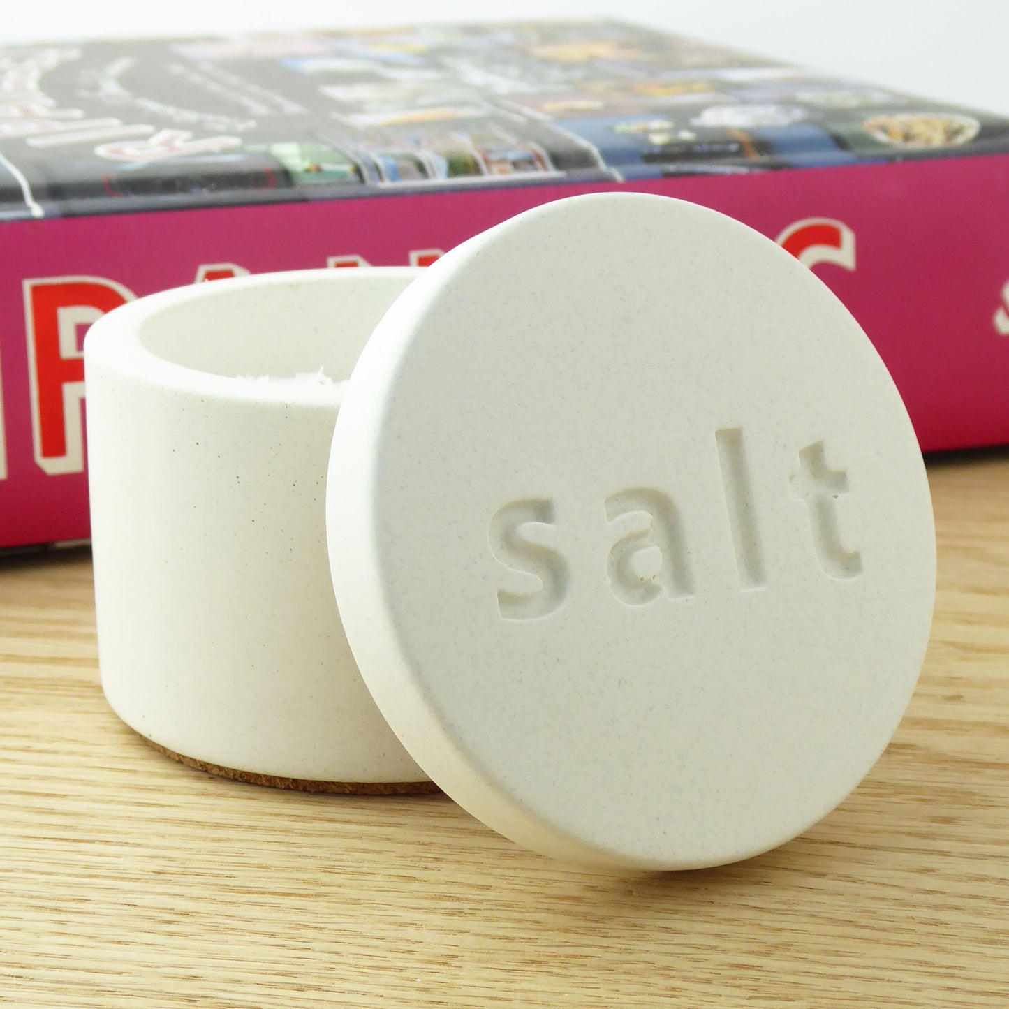 White concrete salt pot with a matching lid with 'salt' engraved on the lid next to a cook book on a shelf