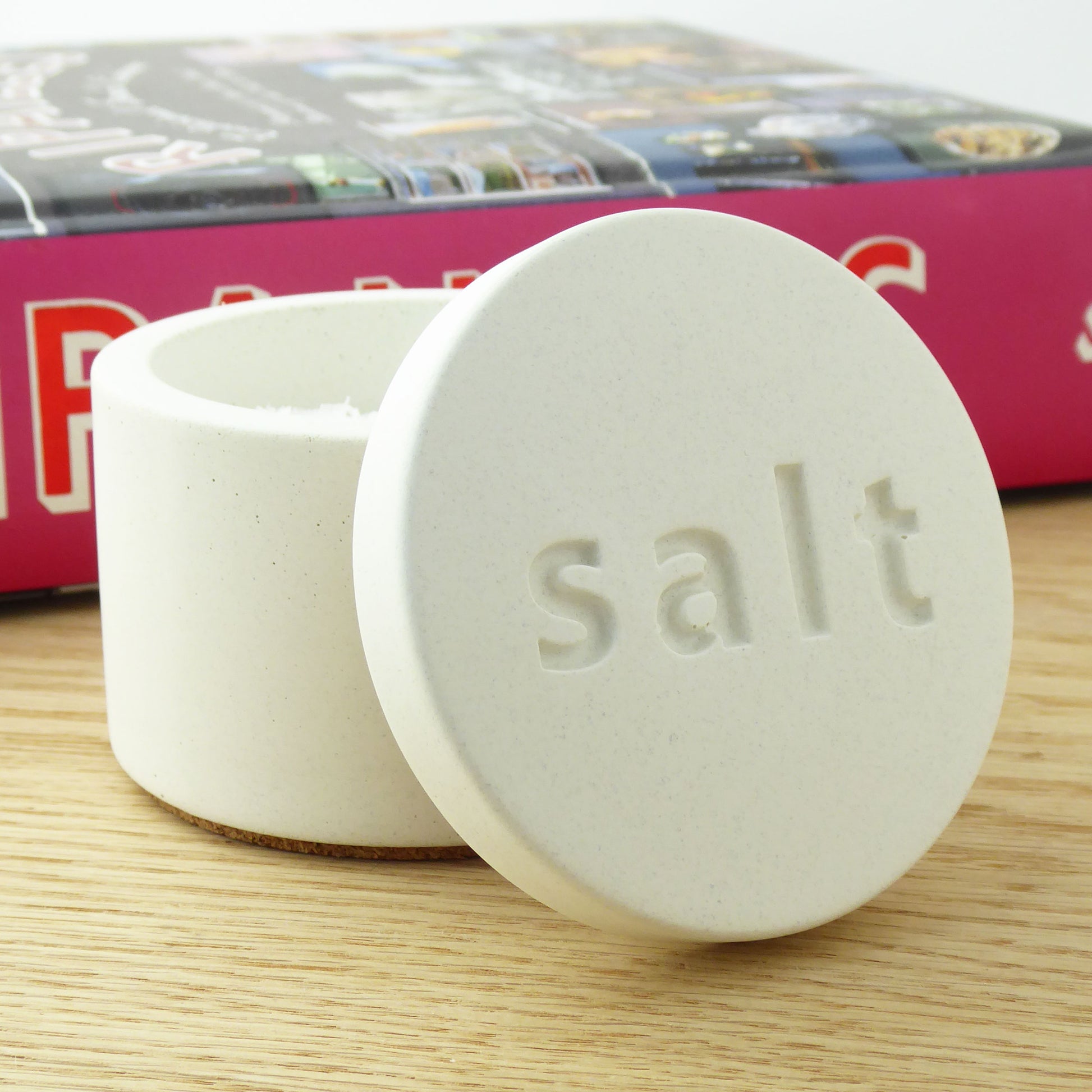 White concrete salt pot with a matching lid with 'salt' engraved on the lid next to a cook book on a shelf