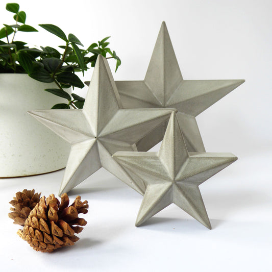 Three star-shaped grey concrete barn star ornaments on a white surface with a plant and festive pine cones in the foreground.