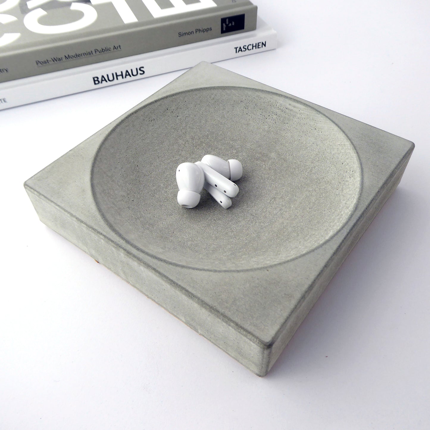 Square grey concrete dish holding ear pods on a white background with books in the background