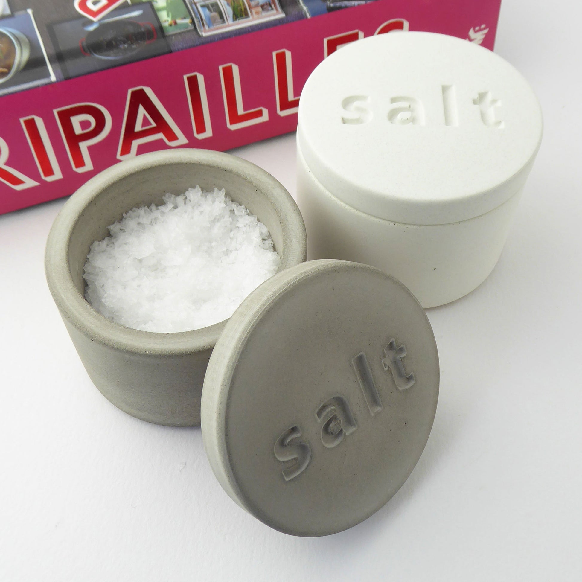 Two concrete salt containers in grey and white with 'salt' engraved on the lids on a white surface, with a background featuring a cookbook.