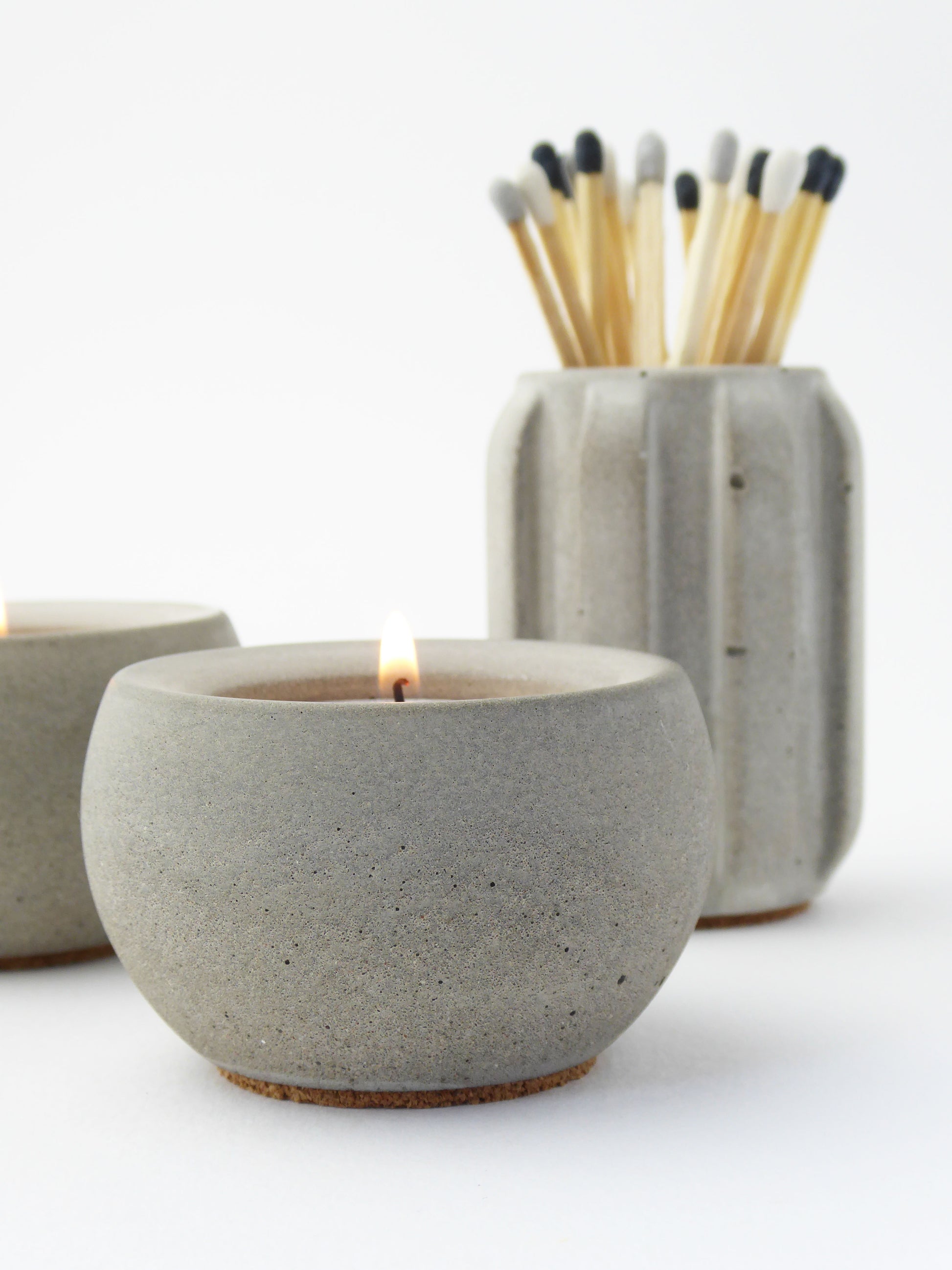Grey concrete candle holder with a lit candle and a grey concrete match pot holding long matches on a white background