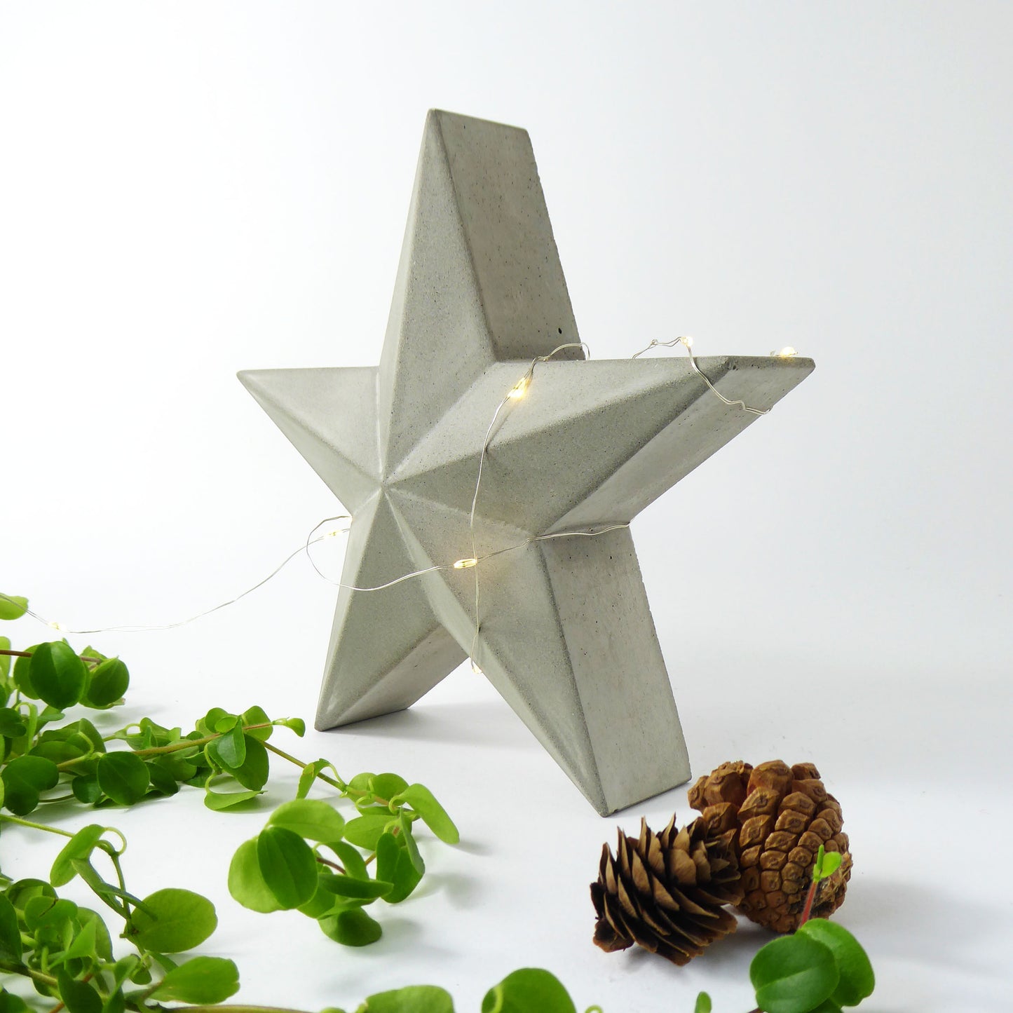 scandi style concrete star with string lights on a white background with greenery and pine cones.