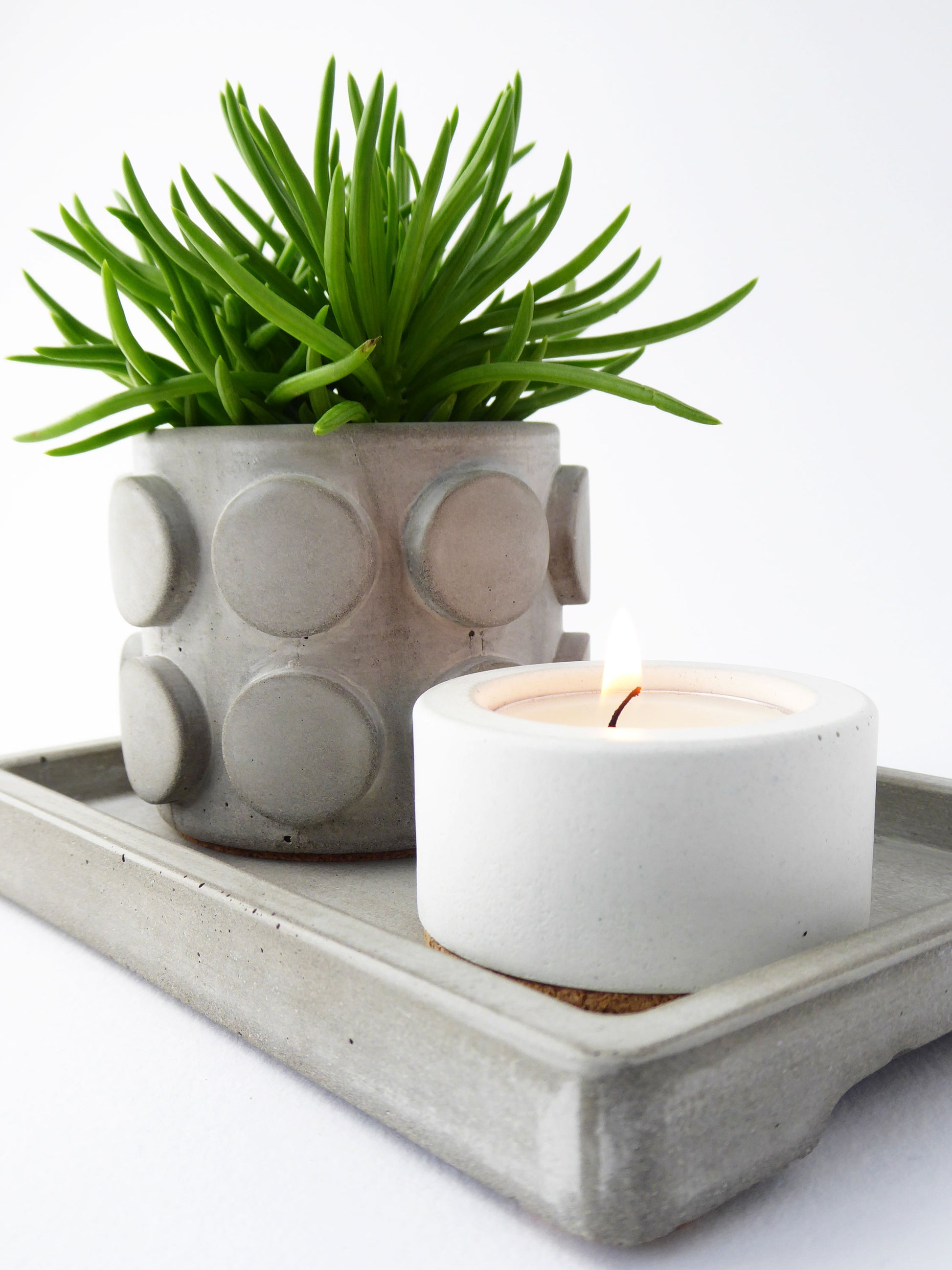 Patterned concrete plant pot with a spiky succulent next to a white candle holder with a lit candle on a grey rectangular concrete tray.