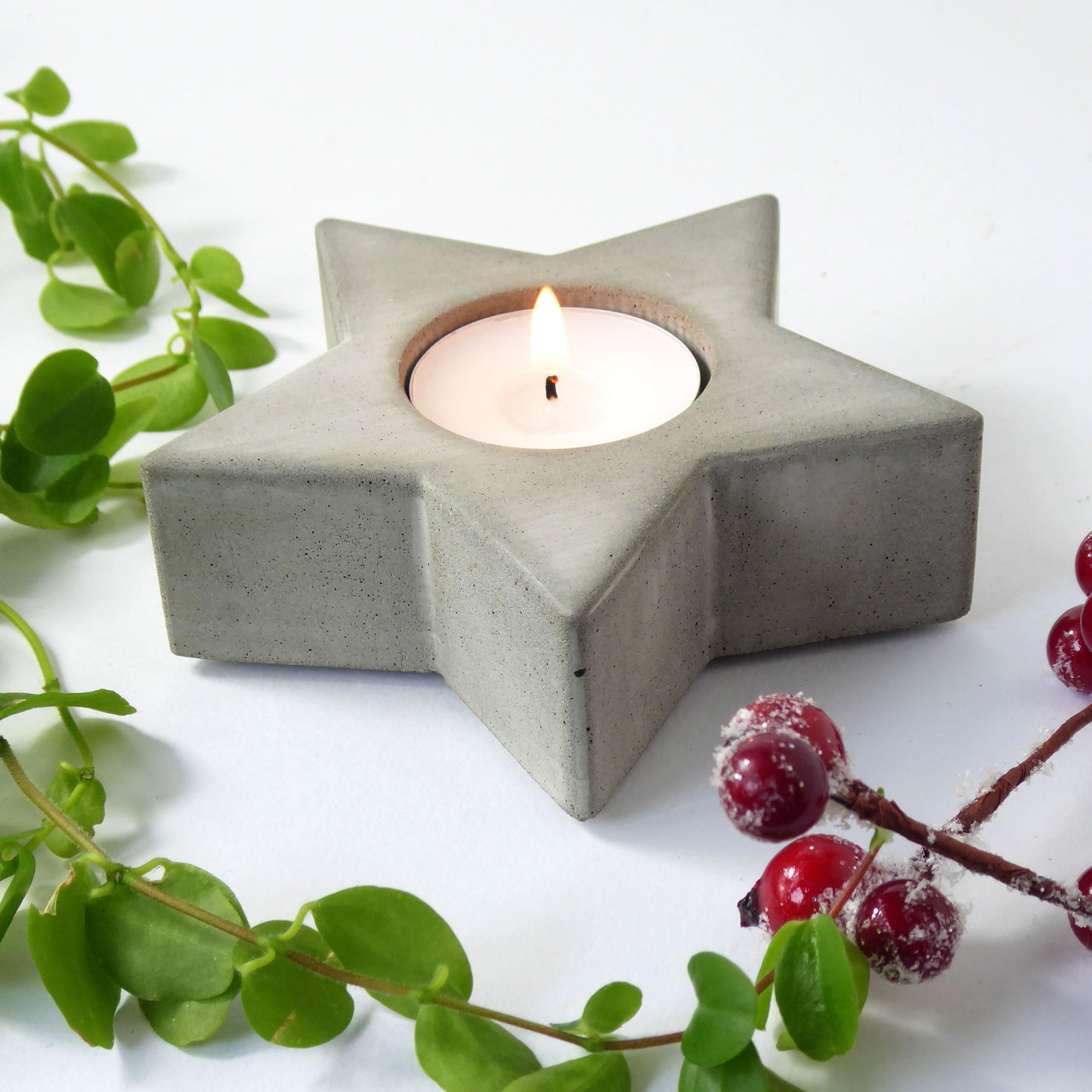 Star-shaped concrete candle holder with a lit candle, surrounded by greenery and Christmas berries on a white background