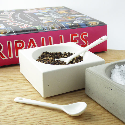 White concrete bowls containing seasalt and black peppercorns with white ceramic spoons on a wooden dining table