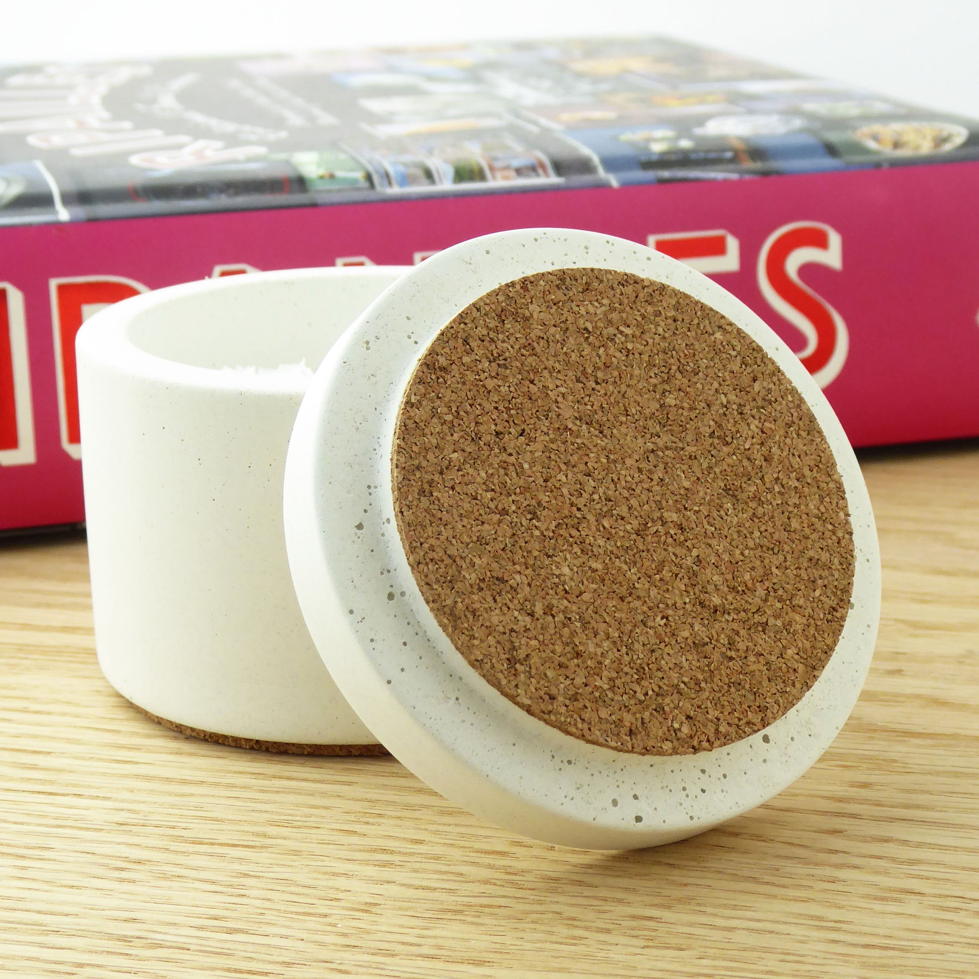 Round concrete pot with a lid with cork on it with a cookbook on a wooden shelf.
