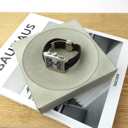 Grey square concrete tray with a men's wristwatch on a beside table