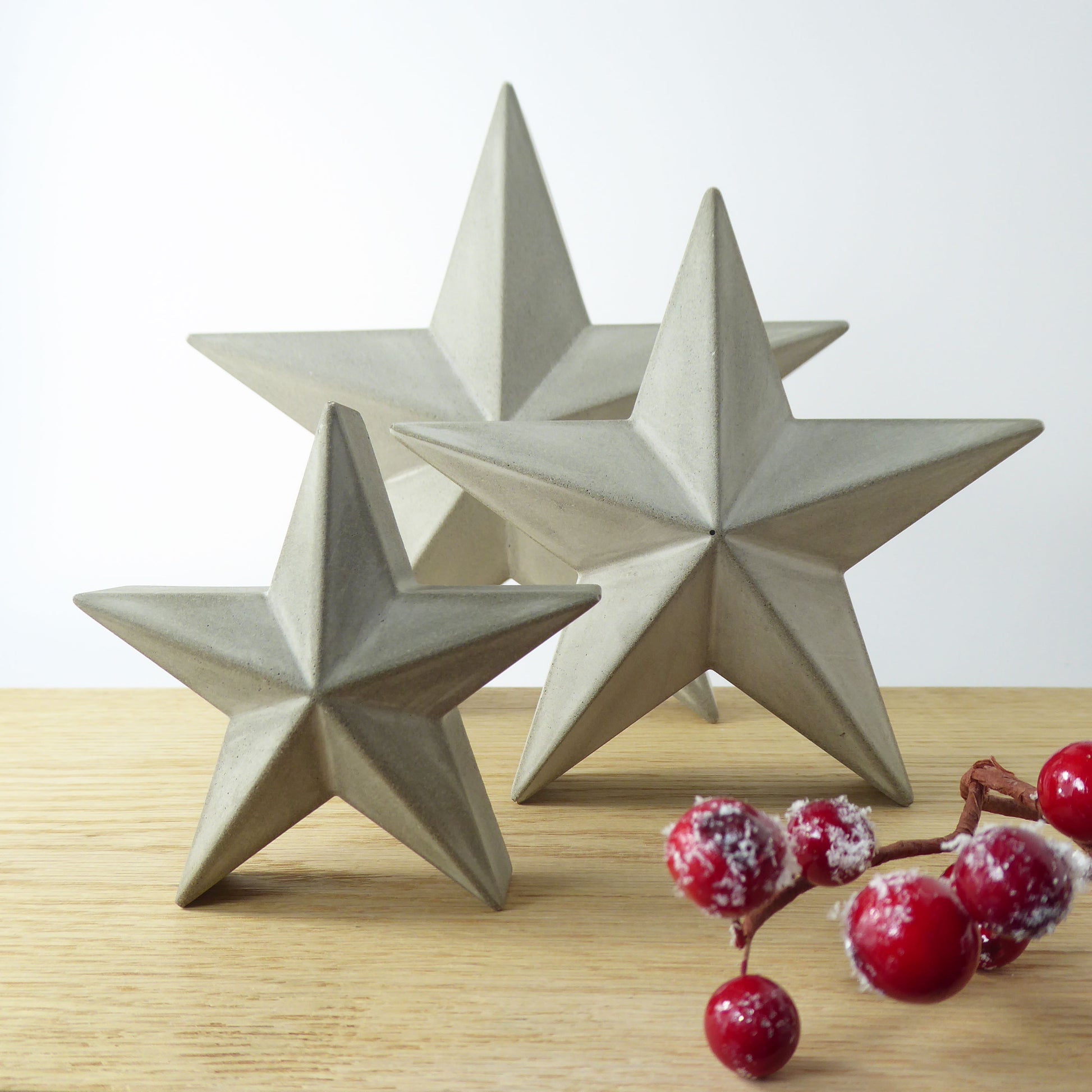 Three star-shaped grey concrete decorative stars on a wooden shelf with festive red berries.