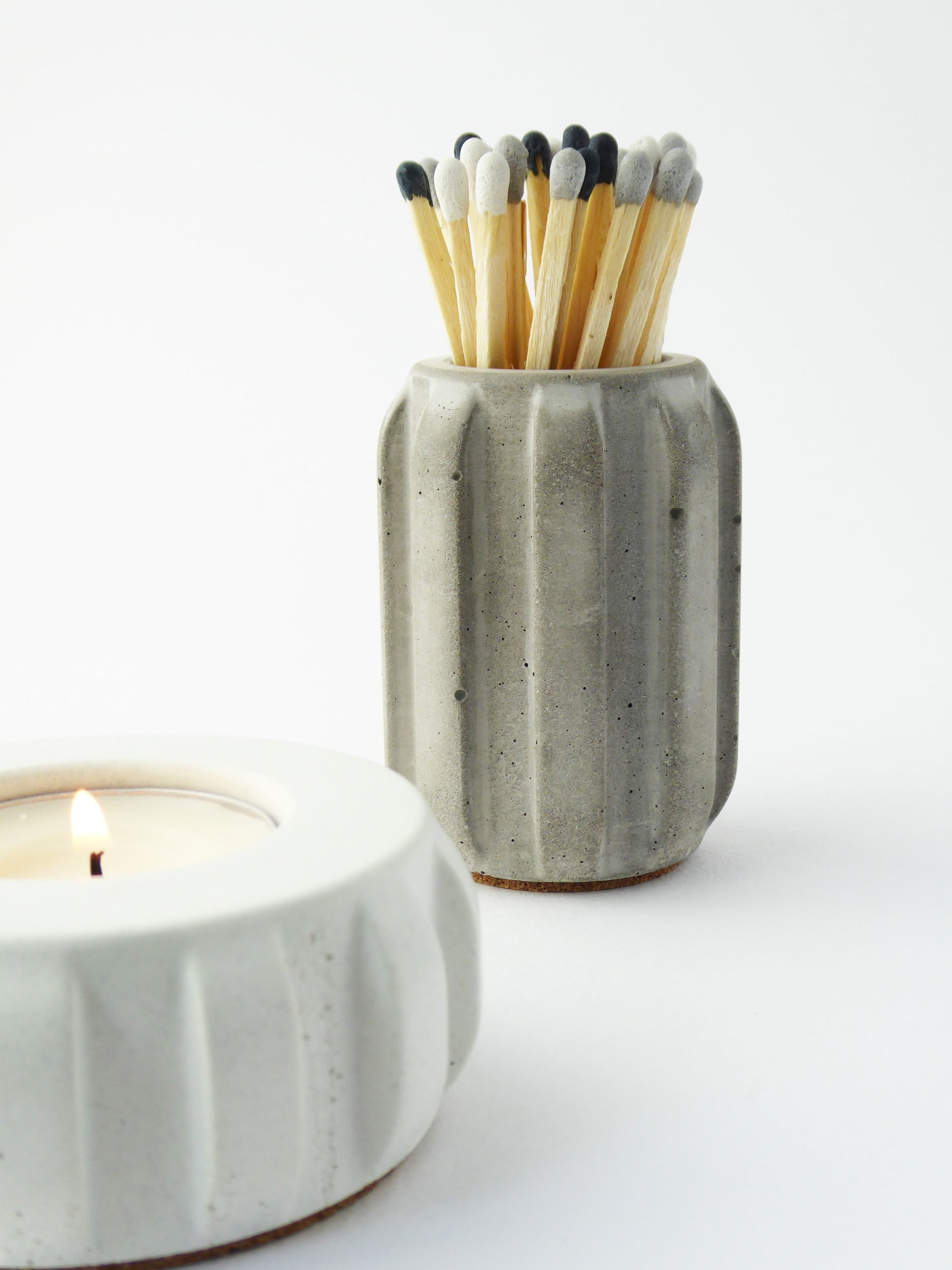 Grey concrete match holder with long coloured matches and a lit candle on a white background