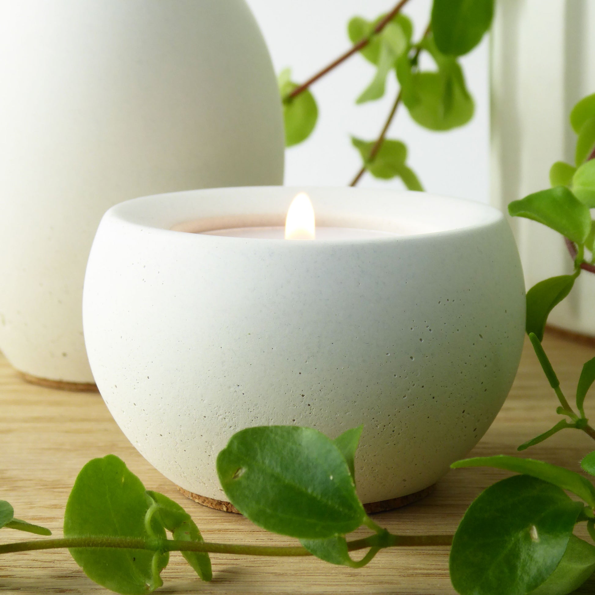 White concrete candle holder with a lit candle on a wooden table with green leaves.