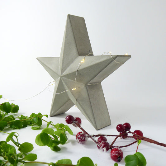 Decorative free standing raw concrete star decoration with string lights on a white background with greenery and festive berries.