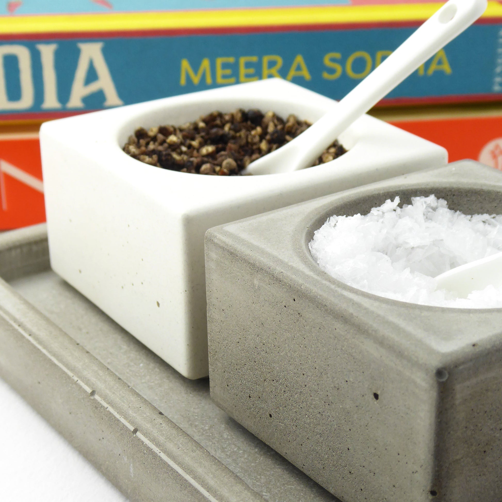 Two white and grey pots with black pepper and seasalt on a grey concrete tray, with a colourful cookbooks in the background.