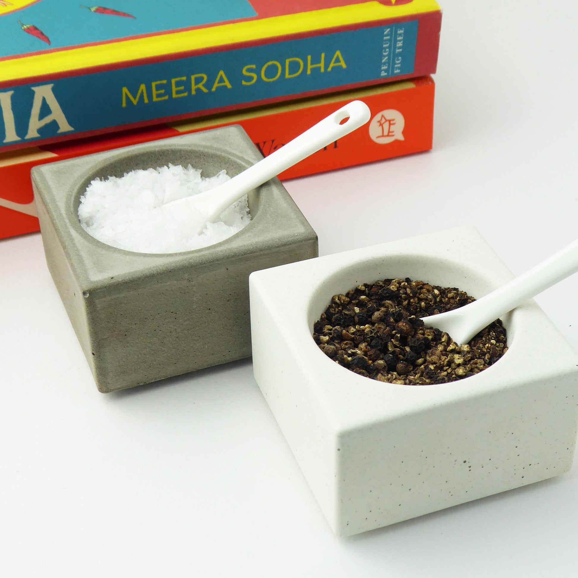 Two concrete bowls with white spoons, one holding salt and the other black peppercorns, on a white surface.