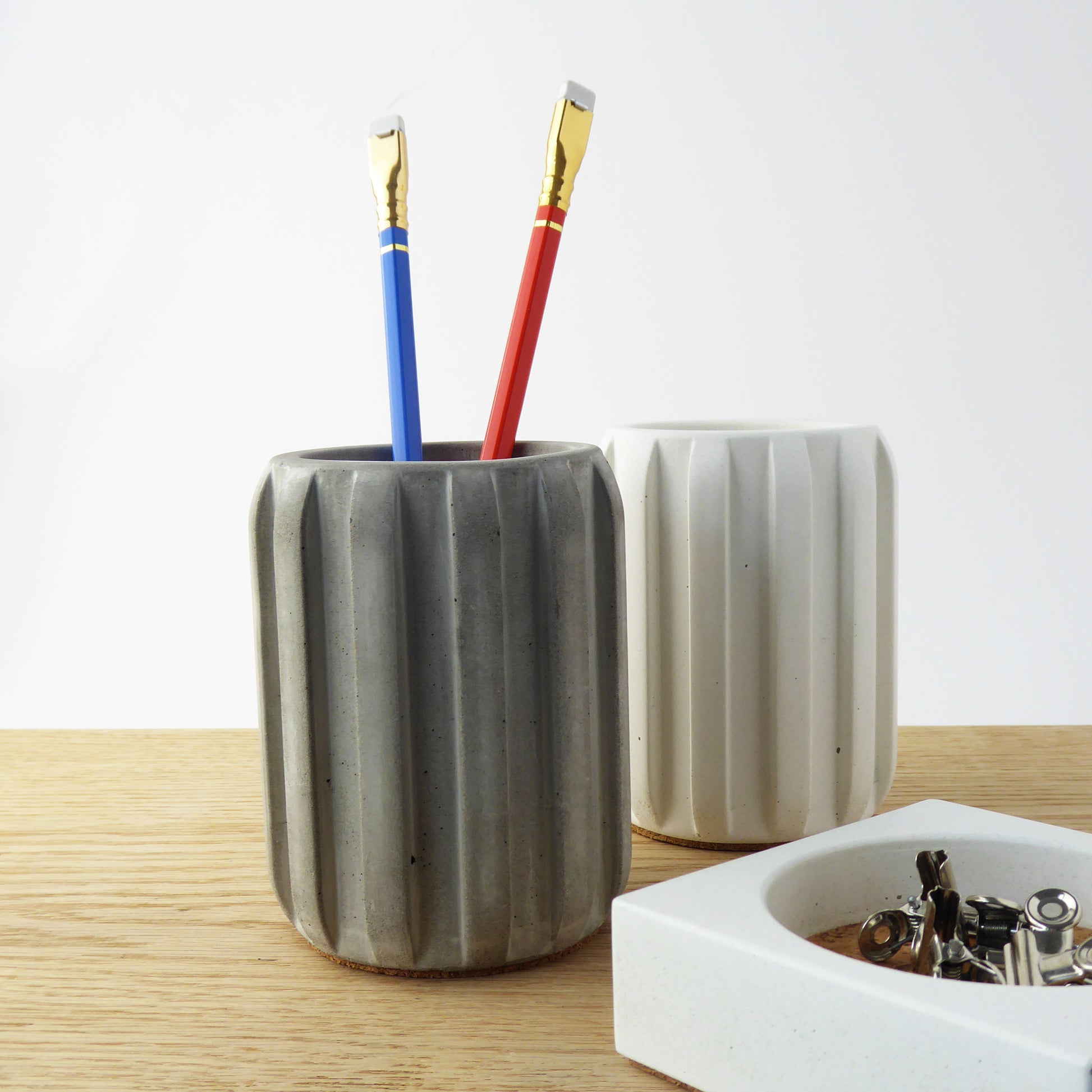Two grey concrete pencil holders with colourful pencils on a wooden surface next to a small concrete bowl containing paperclips.