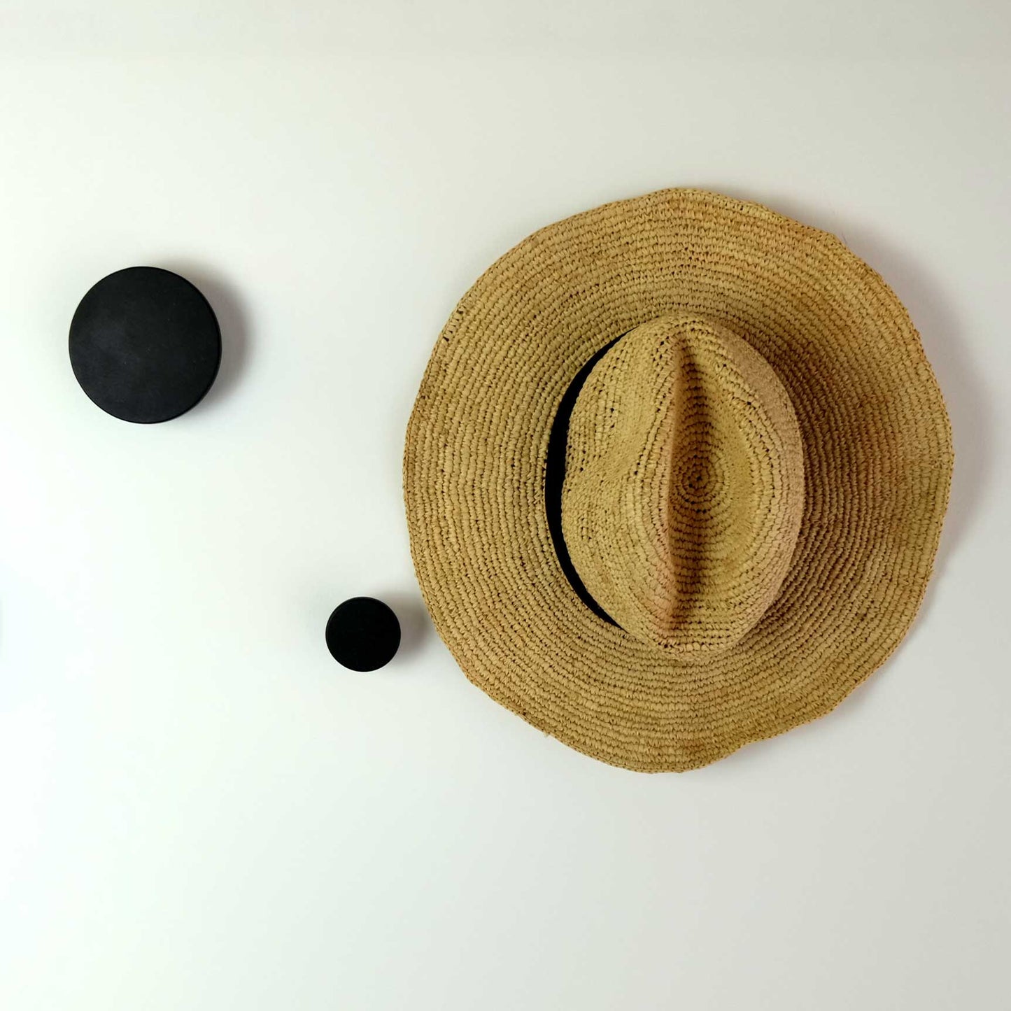 Round black wooden wall hooks of varying sizes mounted on a white wall and one of the hooks has a straw hat hung from it.