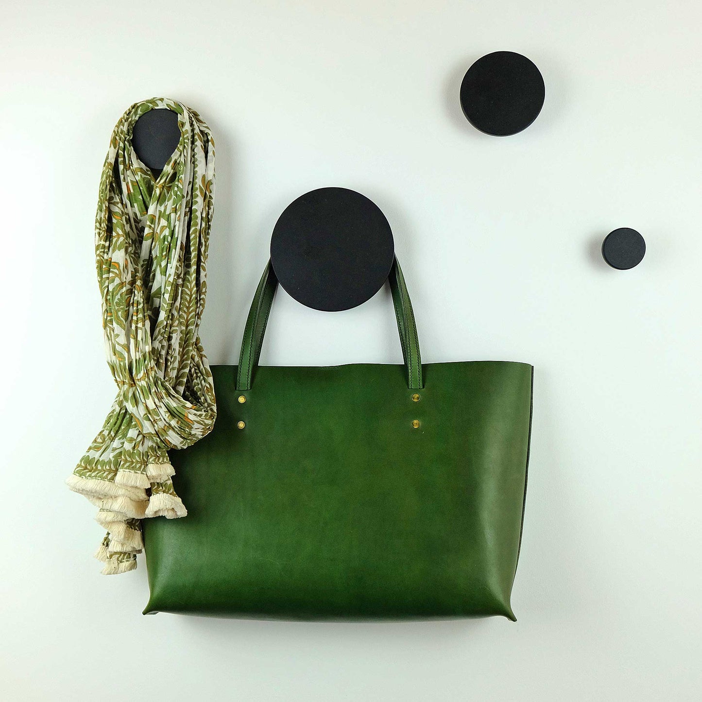 Handmade wooden coat hooks in black in various sizes mounted on a wall with a green leather handbag and a floral print scarf hanging from them