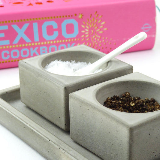 Grey concrete salt and pepper pots and a white ceramic spoon with a pink cookbook in the background