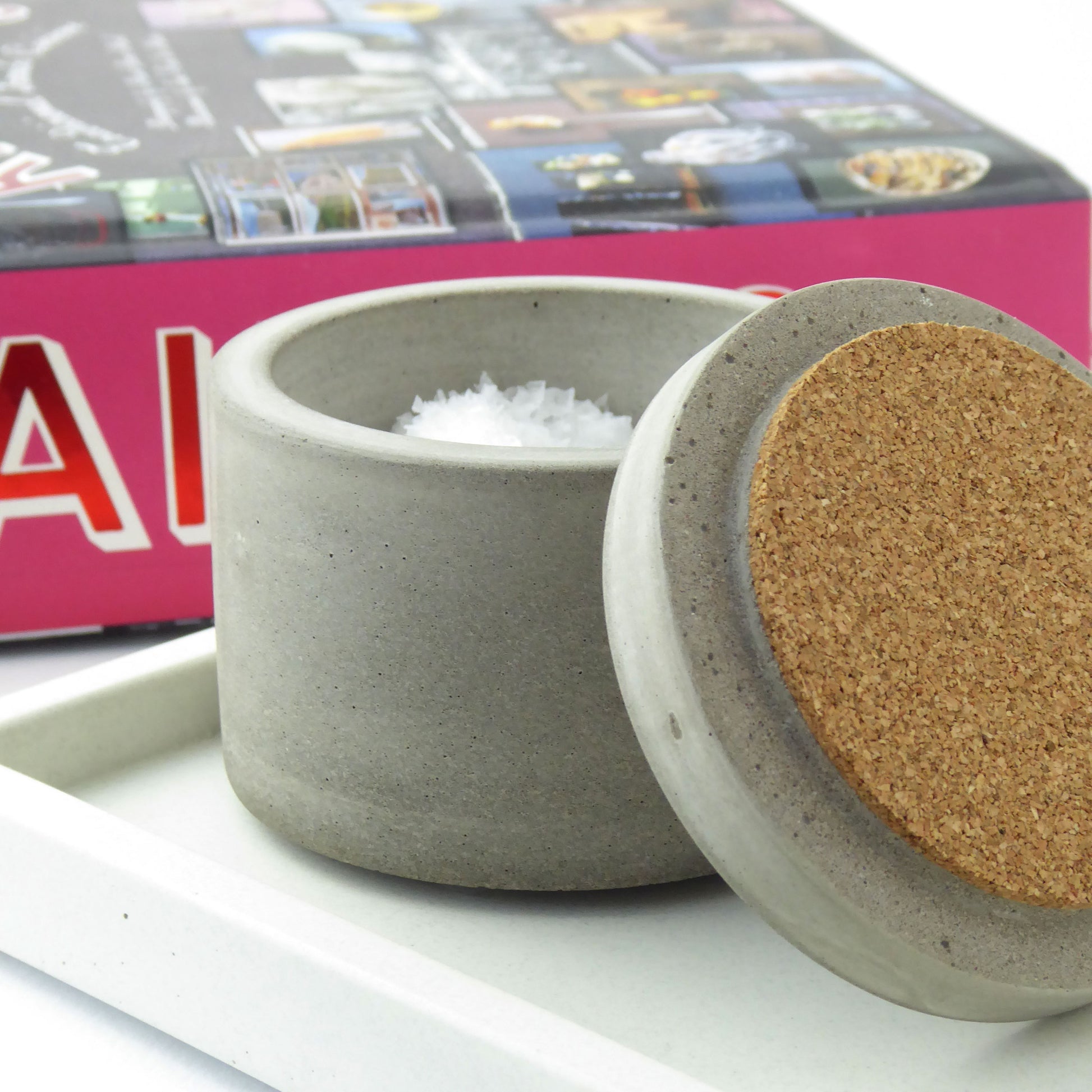 Grey concrete container with salt in it with a concrete and cork lid with a cookbook in the background