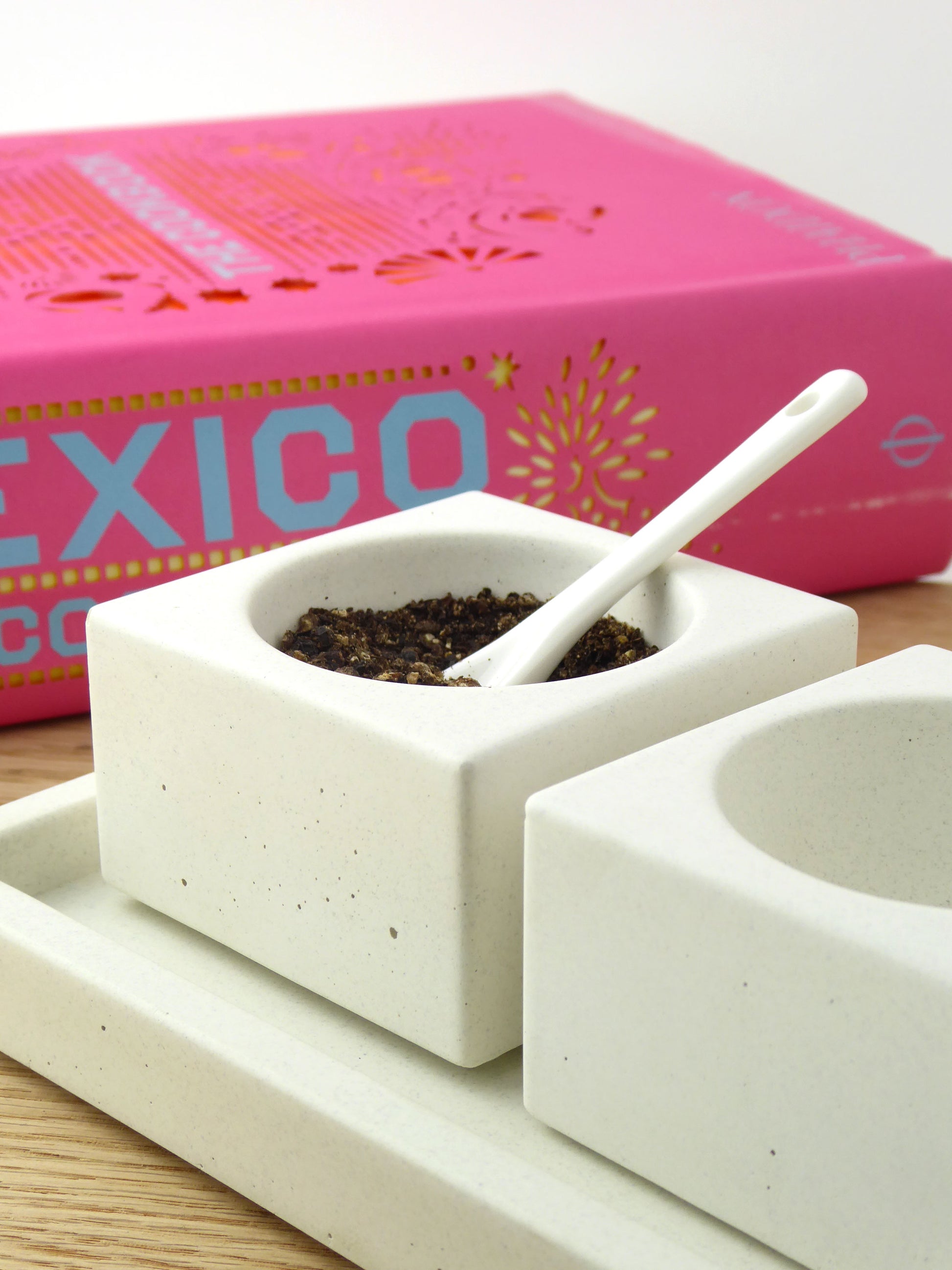White concrete bowls with black peppercorns in one with a white ceramic spoon on a wooden surface, with a pink cookbook
