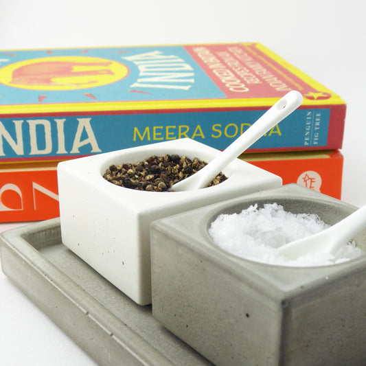Grey and white concrete salt and pepper bowls and white ceramic spoons with cookbooks in the background