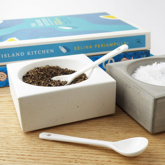 Two concrete bowls with black peppercorns and seasalt and two white spoons on a wooden dining table