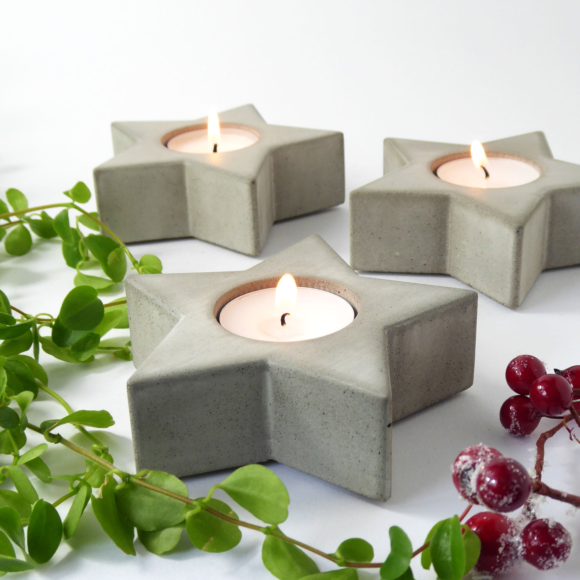 Set of 3 star-shaped concrete candle holders with lit candles on a white surface with greenery and festive berries.