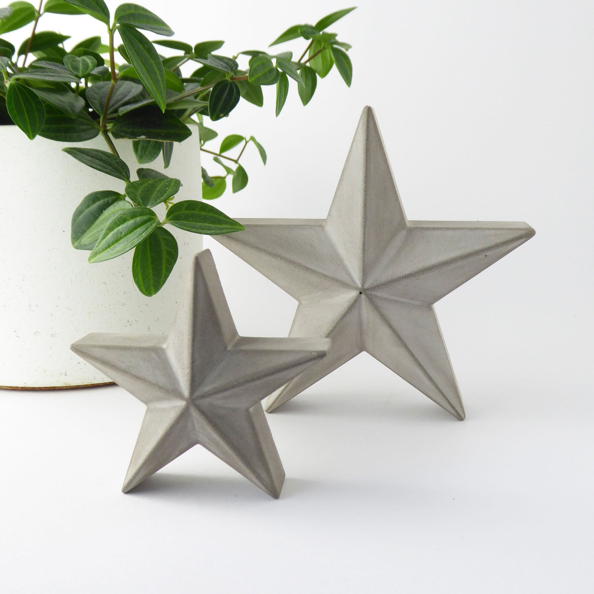 Two concrete star-shaped decorative freestanding ornaments on a white surface with a plant in the background.