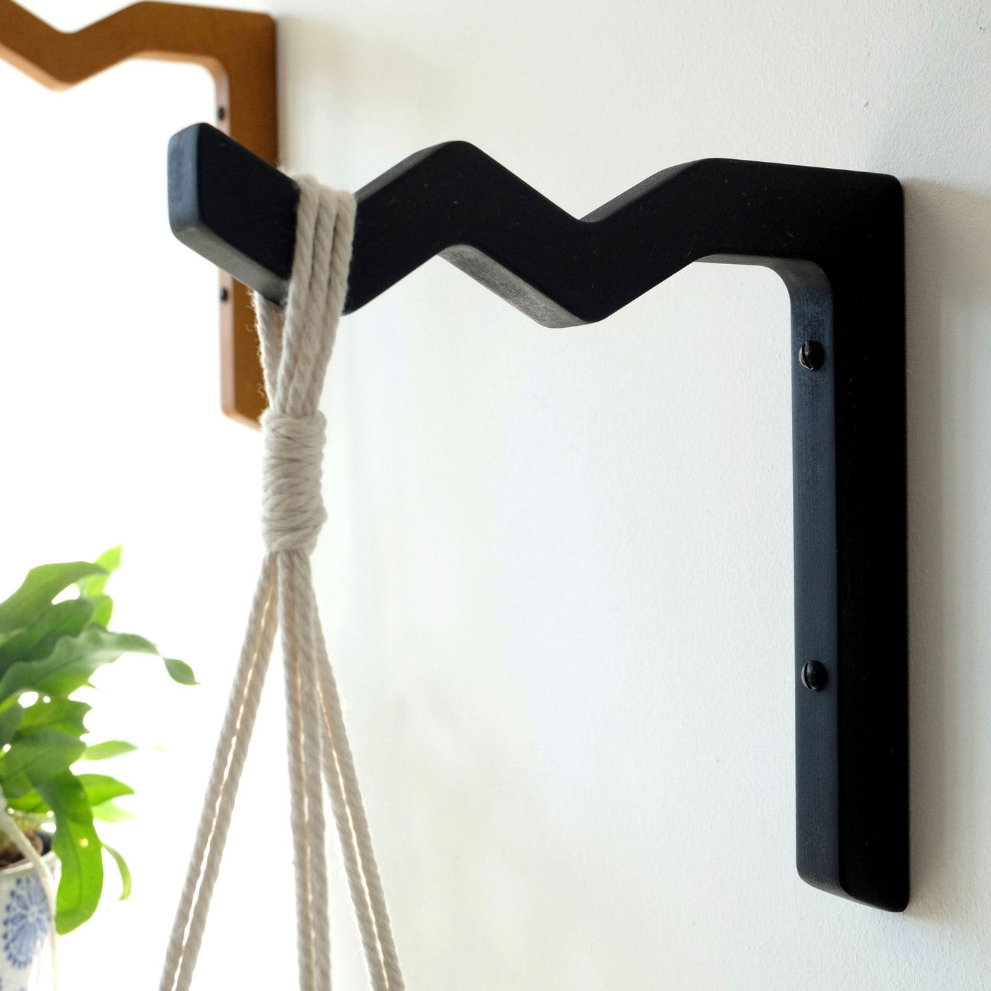 A black wooden indoor wall mounted bracket, mounted on a white wall with a macrame hanger and plant pot hanging from it.