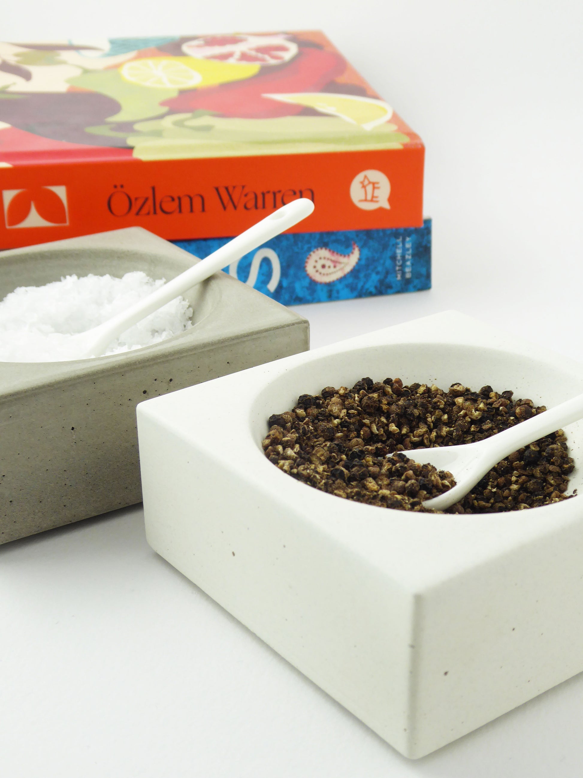 White concrete bowl with black peppercorns and a white spoon, next to a grey concrete bowl with seasalt and a white spoon with colourful cookbooks in the background
