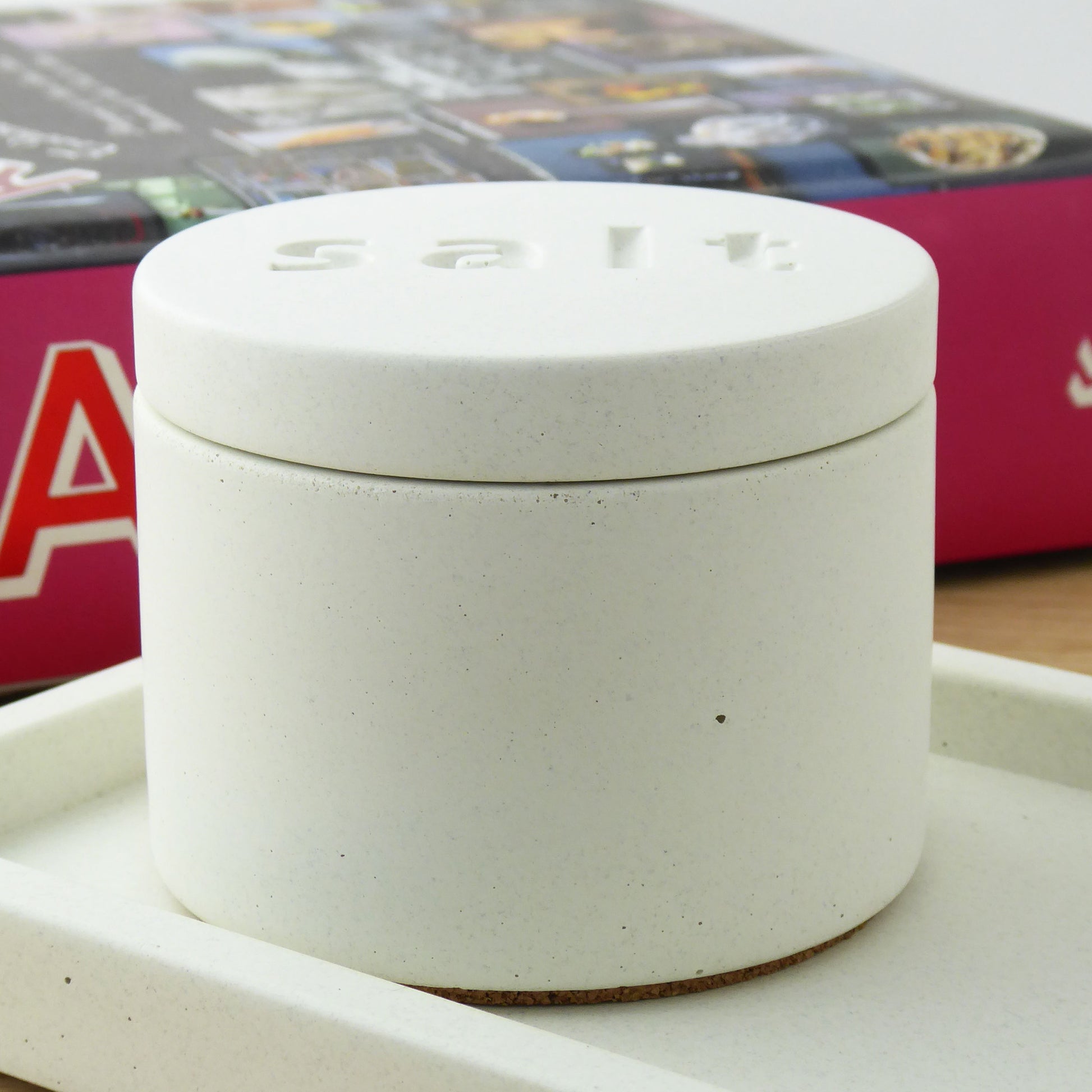 White concrete pot with the words 'salt' engraved on the lid on a white concrete tray with a cookbook in the background