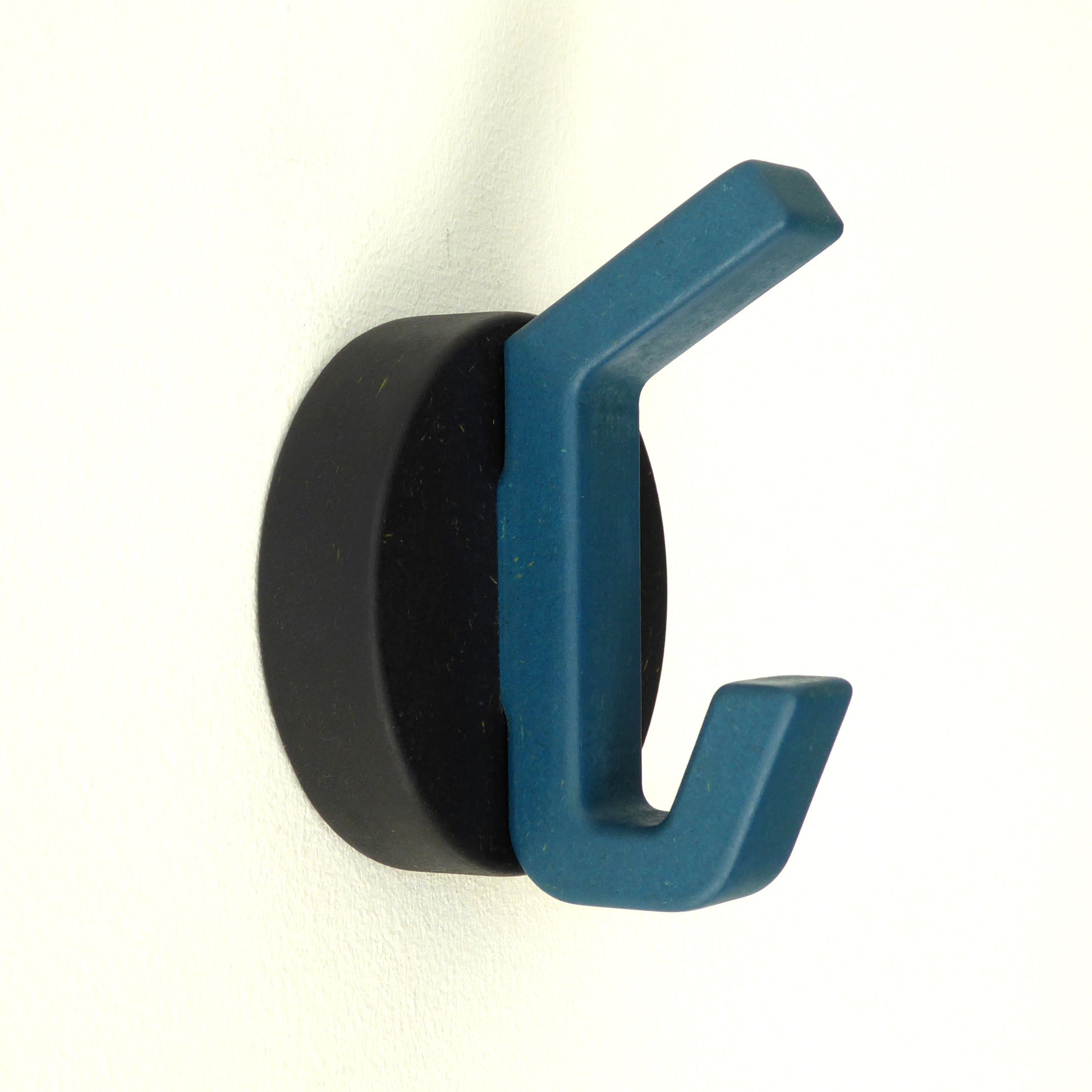 A modern handmade wooden clothes hook with a blue hook on a black round backplate, mounted on a wall.