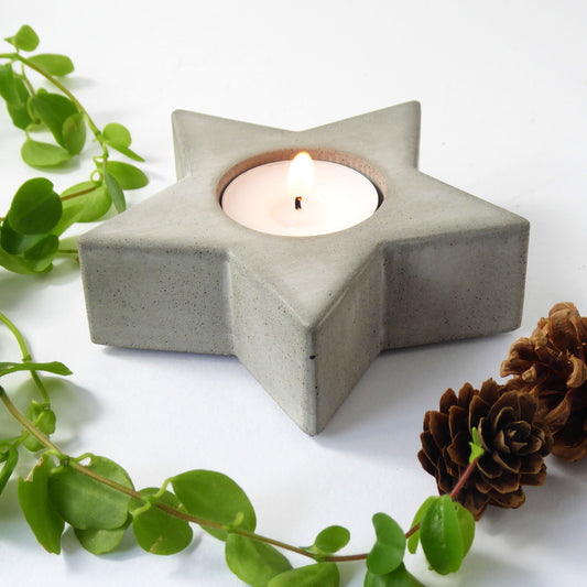 Concrete star-shaped candle holder with a lit candle, surrounded by greenery and pine cones on a white background.
