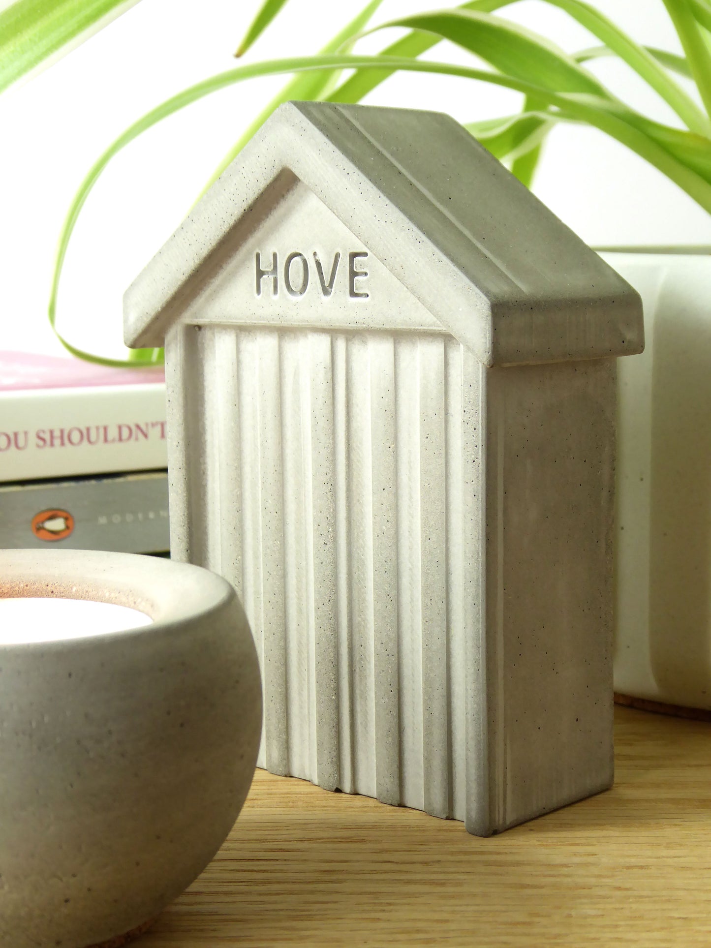 Small concrete beach hut-shaped ornament with 'HOVE' embossed in the design styled on a wooden shelf with books and a houseplant in the background.