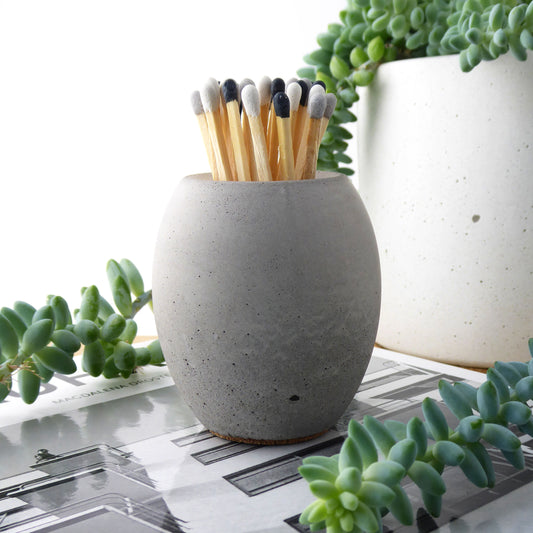 Long matches in a grey concrete match pot holder with green plants in the background