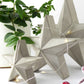 Set of 3 geometric concrete star-shaped freestanding decorations with festive string lights on a white surface with greenery.