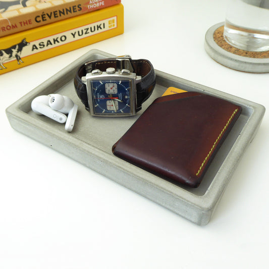 Watch, wallet, and earbuds on a grey concrete rectangular tray with some books styled on a bedside table