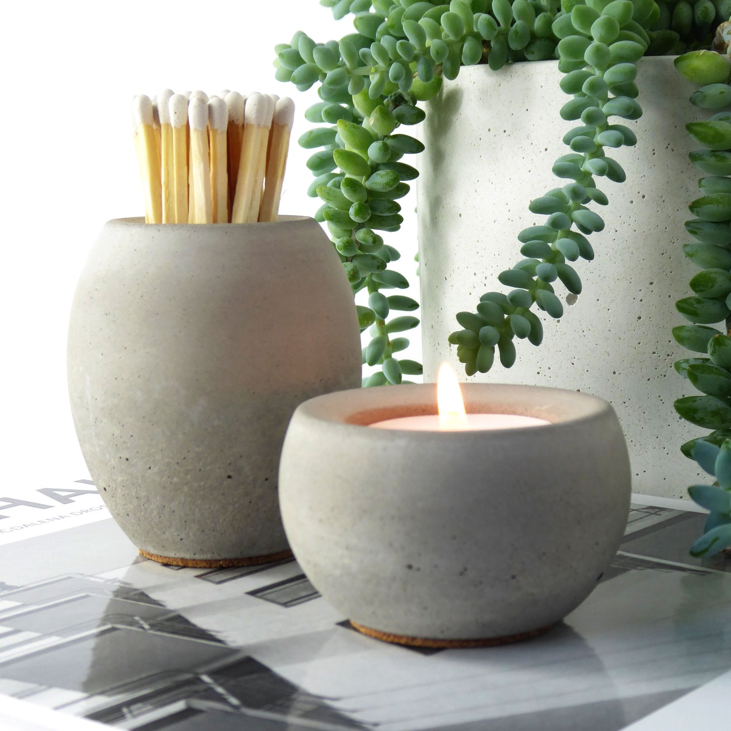 Raw grey concrete match pot holding long matches with a matching grey tea light candle holder positioned next to a succulent plant.