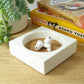 White concrete trinket tray with ear buds inside on a wooden shelf with books and a plant in the background.